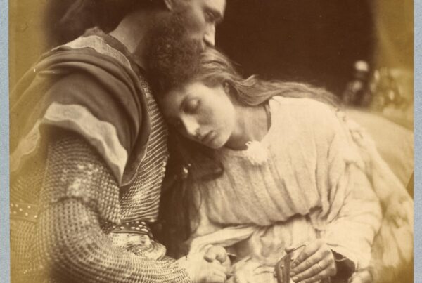 Black and white photograph. Lancelot, dressed in chainmail, holds his lover Guinevere, who leans her head against his chest. She is dressed in a pale coloured smock dress.