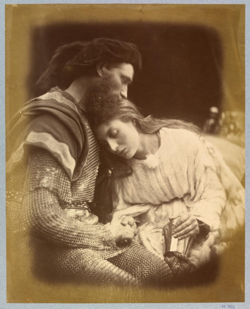 Black and white photograph. Lancelot, dressed in chainmail, holds his lover Guinevere, who leans her head against his chest. She is dressed in a pale coloured smock dress.