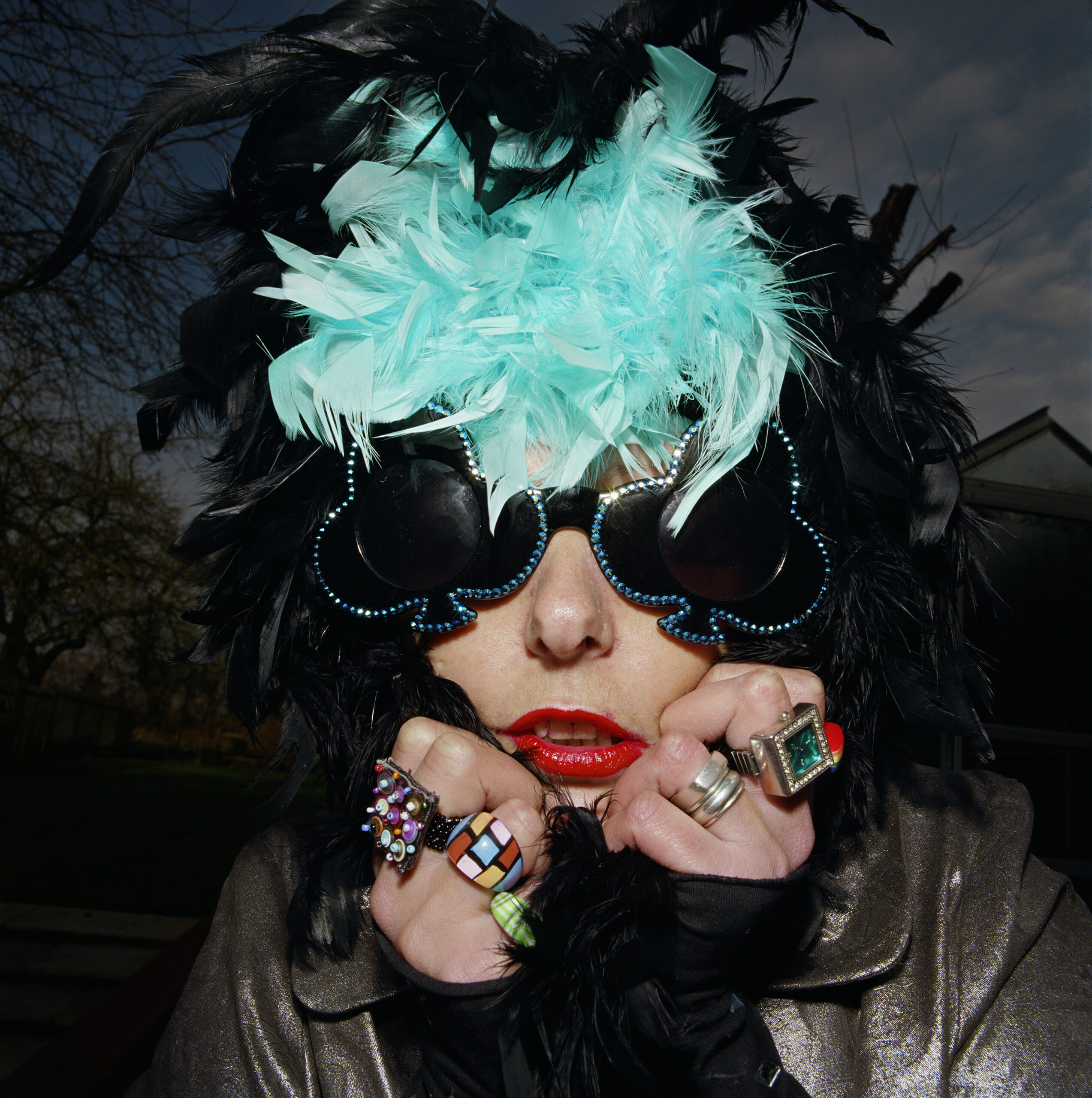 From the exhibition of A Picture of Health, a woman with big jazzy sunglasses is clunching a black and blue feather boa around her head, she's wearing large novelty rings.