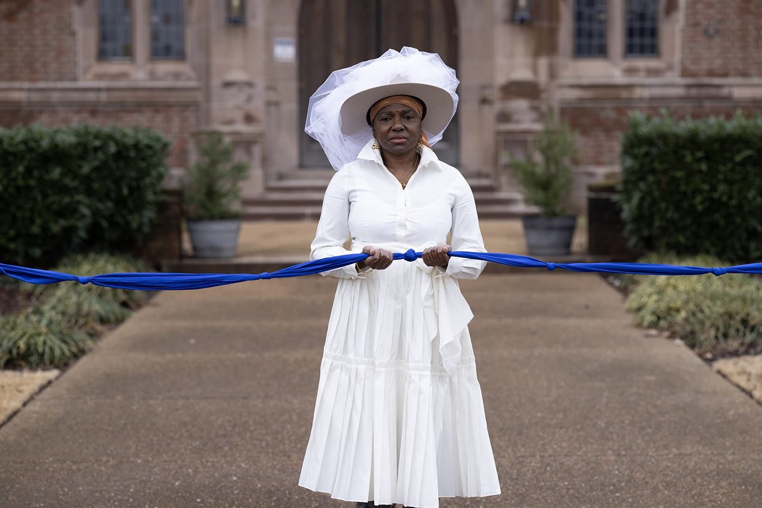 Creator of When We Gather, photographer María Magdalena Campos-Pons standing dressed in white with white hat holding a knotted blue fabric.