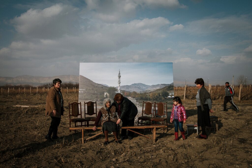 A image of 6 people ranging from a young child to an old woman, in a rural setting. There are mountains in the distant background and in the foreground, the old woman is seated on a bench positioned in front of 4 chairs and a large image depicting a similar landscape.