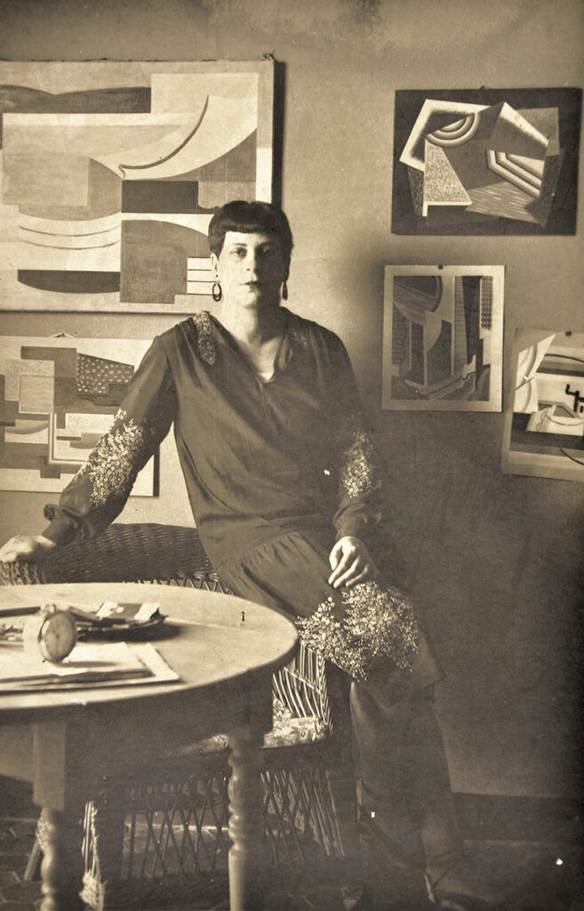 Black and white / sepia toned image. Portrait of Florence Henri leaning against an armchair in her studio.