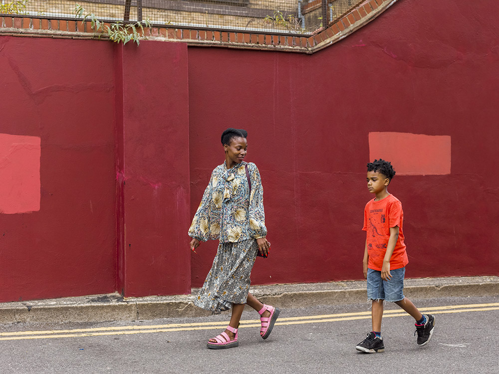 A woman walking slightly ahead of a boy aged around 10. She is smiling at him, while he looks a bit disgruntled. They are walking on a road in front of a bricked wall, painted red, with two patches of lighter red coloured paint. She is stylishly dressed in a long skirt and loose jacket, with fabulous pink, wedged heeled sandals. The boy is wearing a bright red t-shirt, denim shorts and dark trainers.