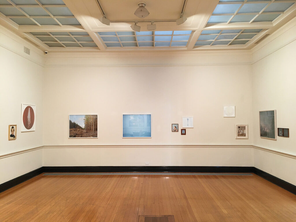 Installation images of Cloudy: A Few Isolated Showers. A light wooden floor, beige walls with scattered artworks along the centra of the image. Glass windows are seen in the ceiling.