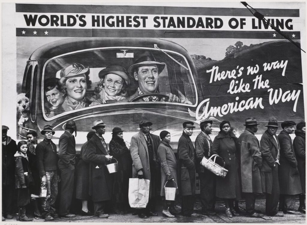 Black and white photograph. People dressed in thick coats and hats queue in front of a painted sign which reads 'World's Highest Standard of Living - There's no way like the American Way.'