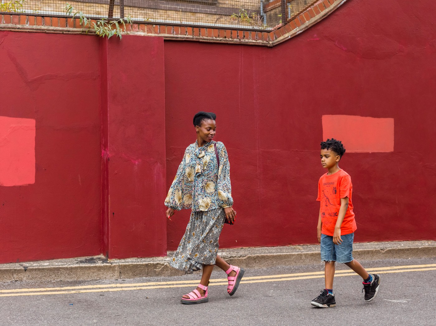 A woman walking slightly ahead of a boy aged around 10. She is smiling at him, while he looks a bit disgruntled. They are walking on a road in front of a bricked wall, painted red, with two patches of lighter red coloured paint. She is stylishly dressed in a long skirt and loose jacket, with fabulous pink, wedged heeled sandals. The boy is wearing a bright red t-shirt, denim shorts and dark trainers.