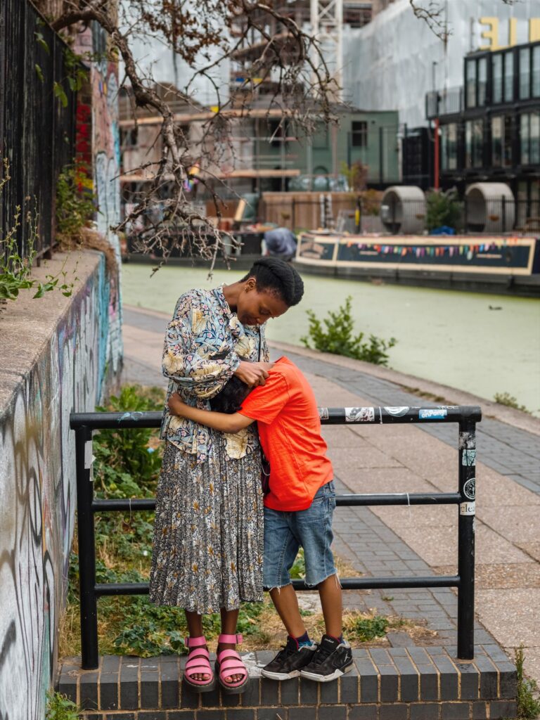 A woman and boy around 10 years of age stood on top of a low brick wall leaning against a three bar, black railing. Behind them is a river running alongside a city scape, with narrow boat in the frame. She is stylishly dressed in a long skirt and loose jacket, with fabulous pink, wedged heeled sandals. The boy is wearing a bright red t shirt, denim shorts and dark trainers. The boy has his arms around the woman in a side hug, his head down and face hidden into her dress. She looks down at him and is stroking his head.