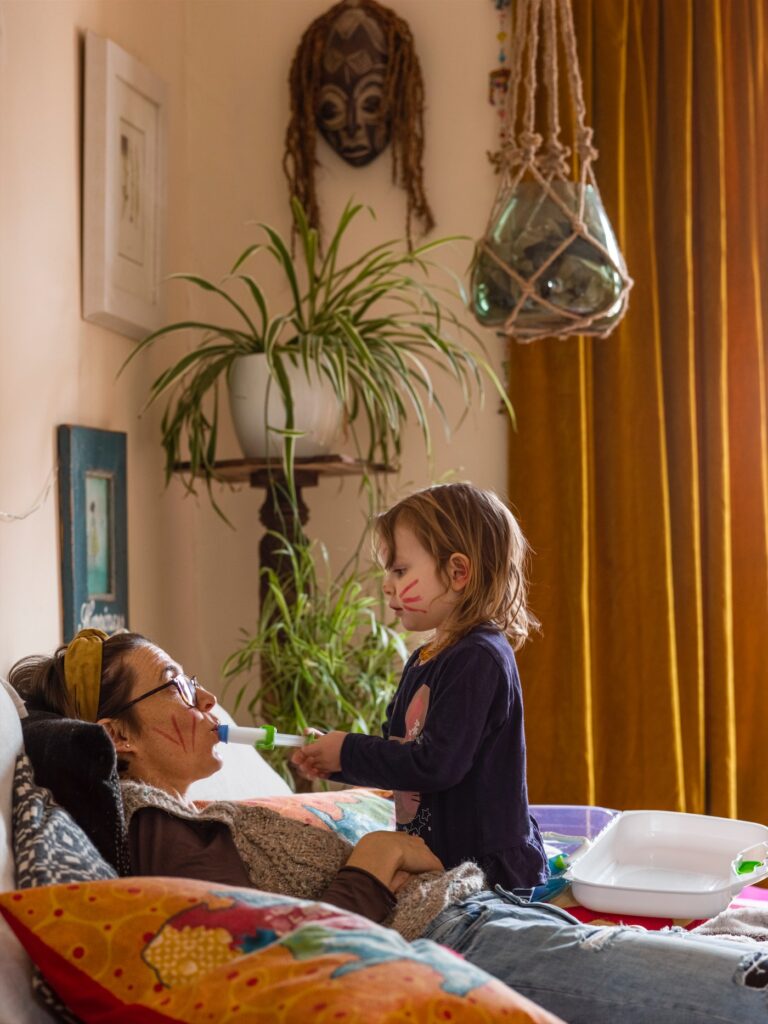 A lounge-type room with amber curtains, a couple of paintings and tribal mask on the wall with huge spider plant on a stand. In the foreground a bespeckled woman, wearing a scarfed hairband, lies against cushions on the sofa apparently playing a game whereby she is a patient being administered medicine into her mouth, via syringe, by her young girl who sits slightly over her. Both have painted red stripes on their faces.