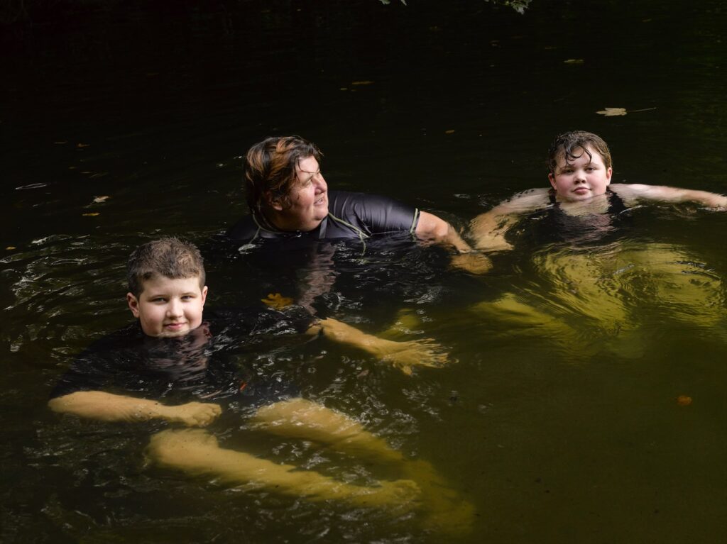 A woman in between two young, teenage children, floating in what appears to be a river. All three are wearing black swimwear, the girl, on the right in a swimming costume, the woman and boy, on the left are wearing t-shirts. Their heads are above the water, outstretched arms and legs, floating out ahead of them under the water. The woman is looking towards the girl while the children are looking up at the photographer.