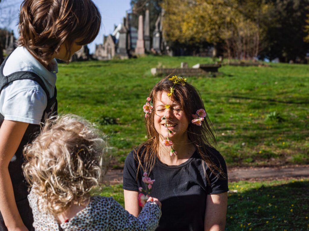 A woman in a kneeling sitting position in front of two children; the one on the right, aged around 7 years looking side on, the younger, around 3 years, a back view. The woman is smiling, eyes shut, sun in her face and adorned with daisy chains around her head and across her face. The background is grassed with graves in the distance on what appears to be a summer’s day.