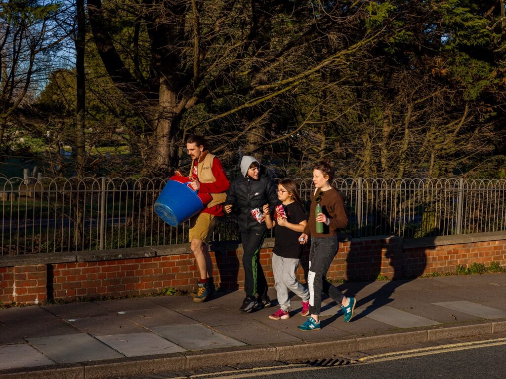 A group of, front left to right, a man, a boy around 13, girl around 11 and a woman, walk along a pavement in front of a railing. The man is carrying a blue truckle. The children are both eating a bag of crisps and looking towards the woman, apparently in conversation. The woman carries a water bottle. All three are dressed for spring/autumn.