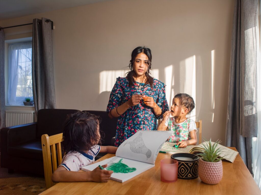 A woman stands next to a table where two children are seated, drawing books open in front of them, crayon/pens in hand, both looking at the woman who is looking wistfully at the photographer.