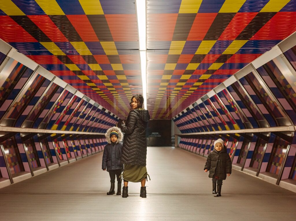 A woman in between two children, the one on the left around 5 years old, the one on the right, slightly further away, around 3 years. They are in a brightly coloured, patterned subway, with red, yellow, blue, black rectangles on the ceiling and the pattern replicated in sunken panels along each wall. All three are wearing winter coats, with the children in hats and black, wellington boots. The children are front facing and look directly at the photographer while the woman, has her back towards the photographer but face turned towards them.