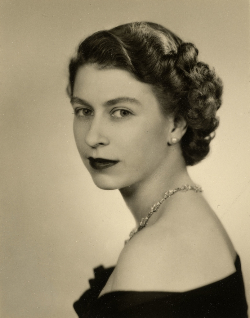 Head and shoulders portrait of HM Queen Elizabeth II, facing the viewer, her torso in left side profile. She wears a black taffeta evening dress with the South Africa Necklace that was a 21st birthday gift from the Government of the Union of South Africa. It was later shortened to fifteen large stones, as shown here, and the remaining diamonds made into a bracelet.