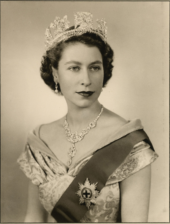 Portrait photograph of HM Queen Elizabeth II, on the occasion of her Accession: a half-length portrait, turned slightly to her left, face. She wears the George IV Diamond Diadem, the Nizam of Hyderabad Necklace, the star and badge of the Order of the Garter on a sash, over a satin evening dress, designed by Norman Hartnell. The Queen is also wearing a diamond and platinum bracelet that was given as a wedding present by Prince Philip. This was designed by Philip Antrobus, using stones taken a from a tiara that had belonged to Prince Philip's mother Princess Andrew of Greece. She is also holding a fan in her left hand.