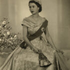 Photograph of HM Queen Elizabeth II, on the occasion of her Accession: a three-quarter length portrait, her face turned to her right. She wears the Diamond Diadem, the Nizam of Hyderabad Necklace, the star of the Order of the Garter on a sash, over a satin evening dress, designed by Norman Hartnell. The Queen is also wearing a diamond and platinum bracelet that was given as a wedding present by Prince Philip. This was designed by Philip Antrobus, using stones taken a from a tiara that had belonged to Prince Philip's mother Princess Andrew of Greece. She is also holding a fan in her left hand. A vase of flowers in background to left.