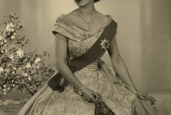 Photograph of HM Queen Elizabeth II, on the occasion of her Accession: a three-quarter length portrait, her face turned to her right. She wears the Diamond Diadem, the Nizam of Hyderabad Necklace, the star of the Order of the Garter on a sash, over a satin evening dress, designed by Norman Hartnell. The Queen is also wearing a diamond and platinum bracelet that was given as a wedding present by Prince Philip. This was designed by Philip Antrobus, using stones taken a from a tiara that had belonged to Prince Philip's mother Princess Andrew of Greece. She is also holding a fan in her left hand. A vase of flowers in background to left.