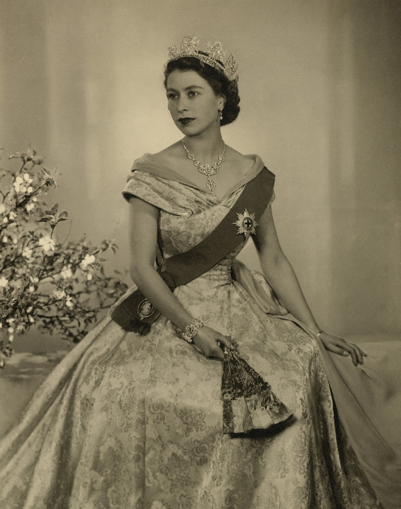 Photograph of HM Queen Elizabeth II, on the occasion of her Accession: a three-quarter length portrait, her face turned to her right. She wears the Diamond Diadem, the Nizam of Hyderabad Necklace, the star of the Order of the Garter on a sash, over a satin evening dress, designed by Norman Hartnell. The Queen is also wearing a diamond and platinum bracelet that was given as a wedding present by Prince Philip. This was designed by Philip Antrobus, using stones taken a from a tiara that had belonged to Prince Philip's mother Princess Andrew of Greece. She is also holding a fan in her left hand. A vase of flowers in background to left.