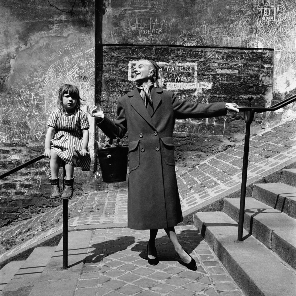 Black and white photo. A child sits on a railing next to woman in a long coat, who stands smiling.