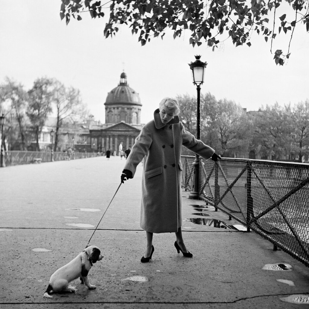 Black and white photo. A woman in a long coat walks her dog on a lead. The dog is seated to the left of her.