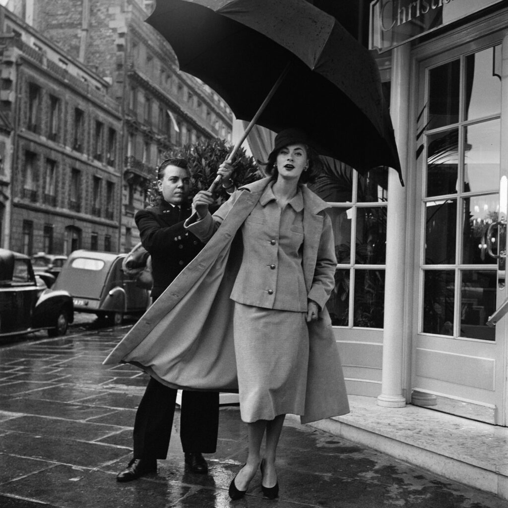 Black and white photo. A man holds an umbrella above a woman's head outside of a shop.