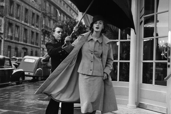 Black and white photo. A man holds an umbrella above a woman's head outside of a shop.