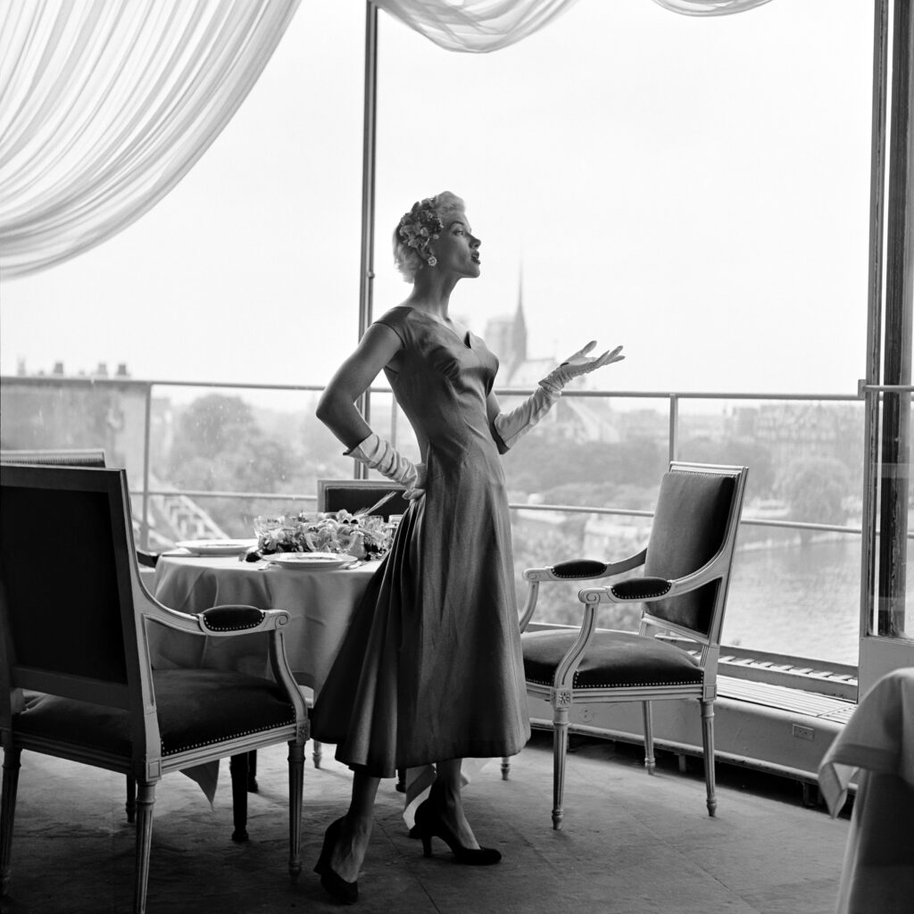 Black and white photo. A woman stands next to a table and two chairs. One of her hands is on her hip, the other is gesturing in front of her.