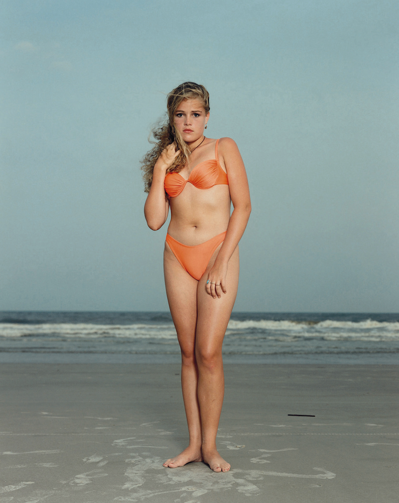 A young woman wears an orange bikini while standing on a beach in front of the sea.