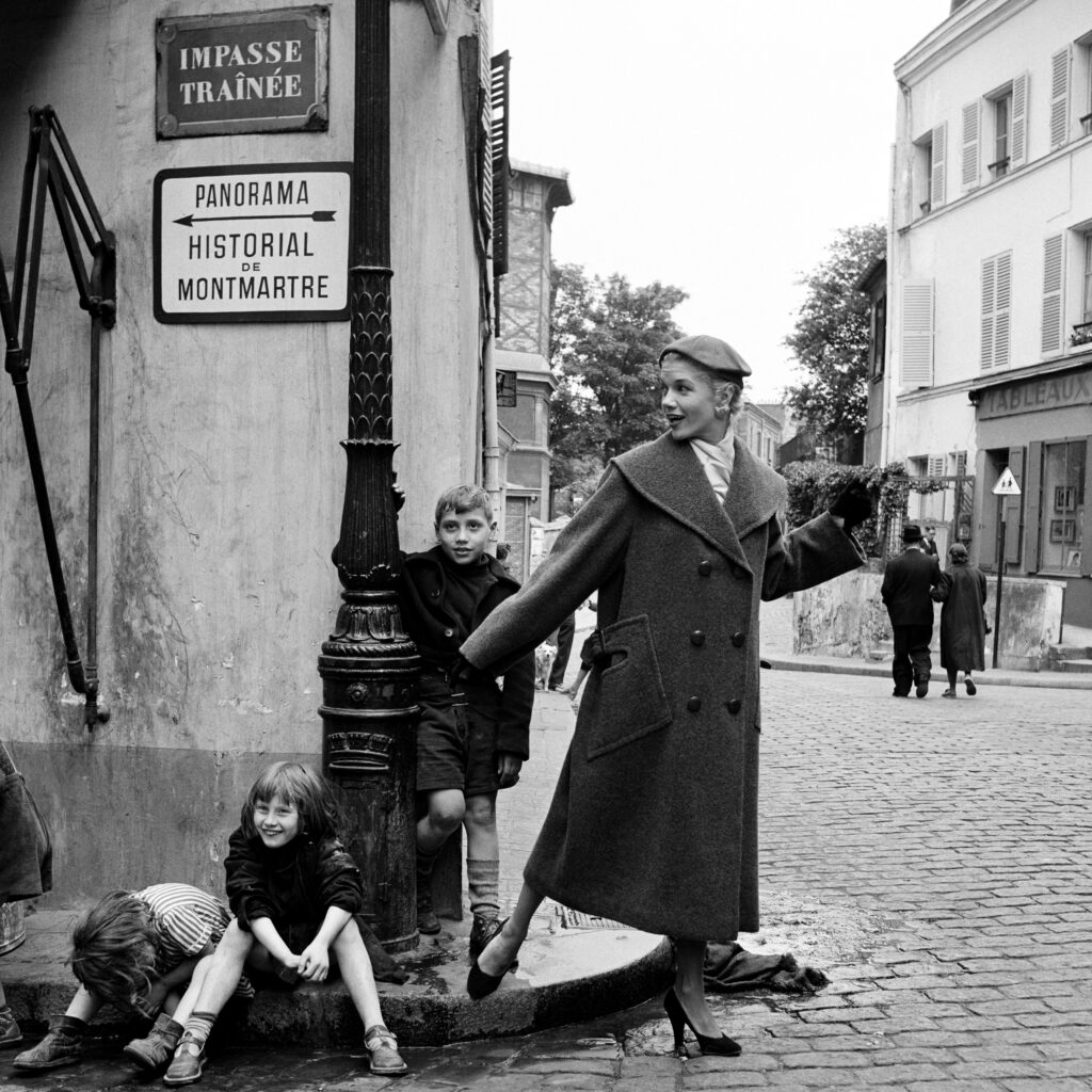 Black and white photo. Two children are seated on a curb while another child and a woman stand beside them.