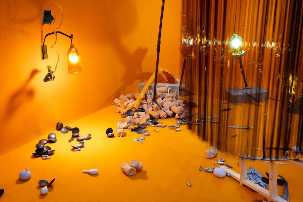Installation image. A room bathed in orange light. Objects resembling concrete drinking cups are piled in one corner, while black streamers hang down nearby.