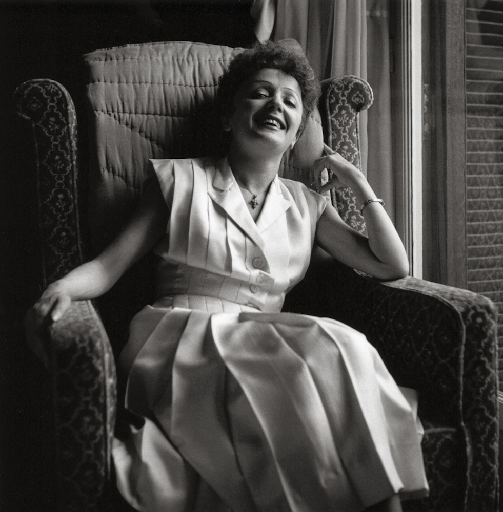 Édith Piaf sits in an armchair, looking towards the camera and smiling.