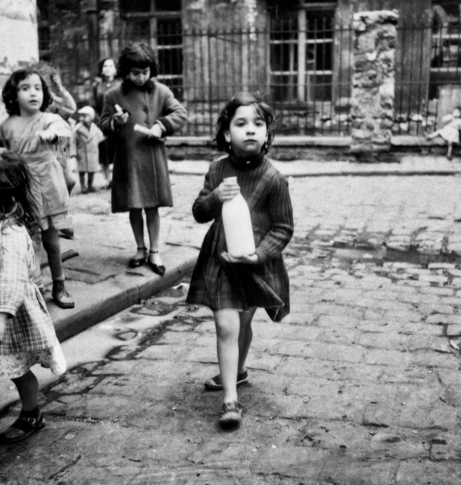 Black and white photo of a young girl carrying a milk bottle. There are other children walking in the background.