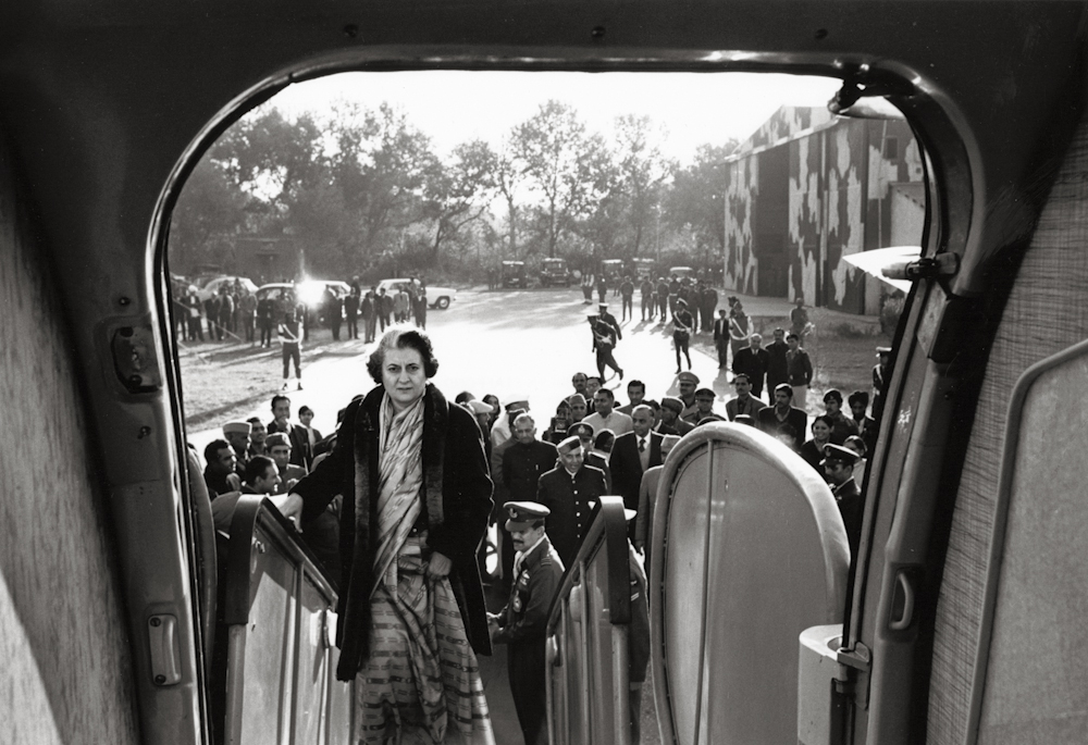 Black and white image taken from a plane. The camera is pointed down the stairs, showing a woman boarding. There is a small crowd gathered behind her.