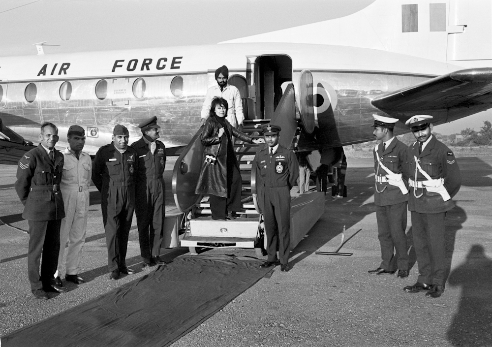 Black and white photo. A woman (Marilyn Stafford) pauses as she walks up stairs leading into a plane. A group of men in various uniforms stand around the plane, looking towards the camera.