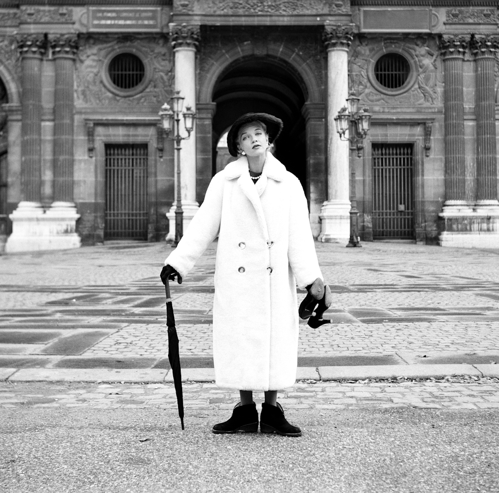 Black and white photograph. A woman in a long coat stands holding an umbrella in one hand and a pair of shoes in the other.