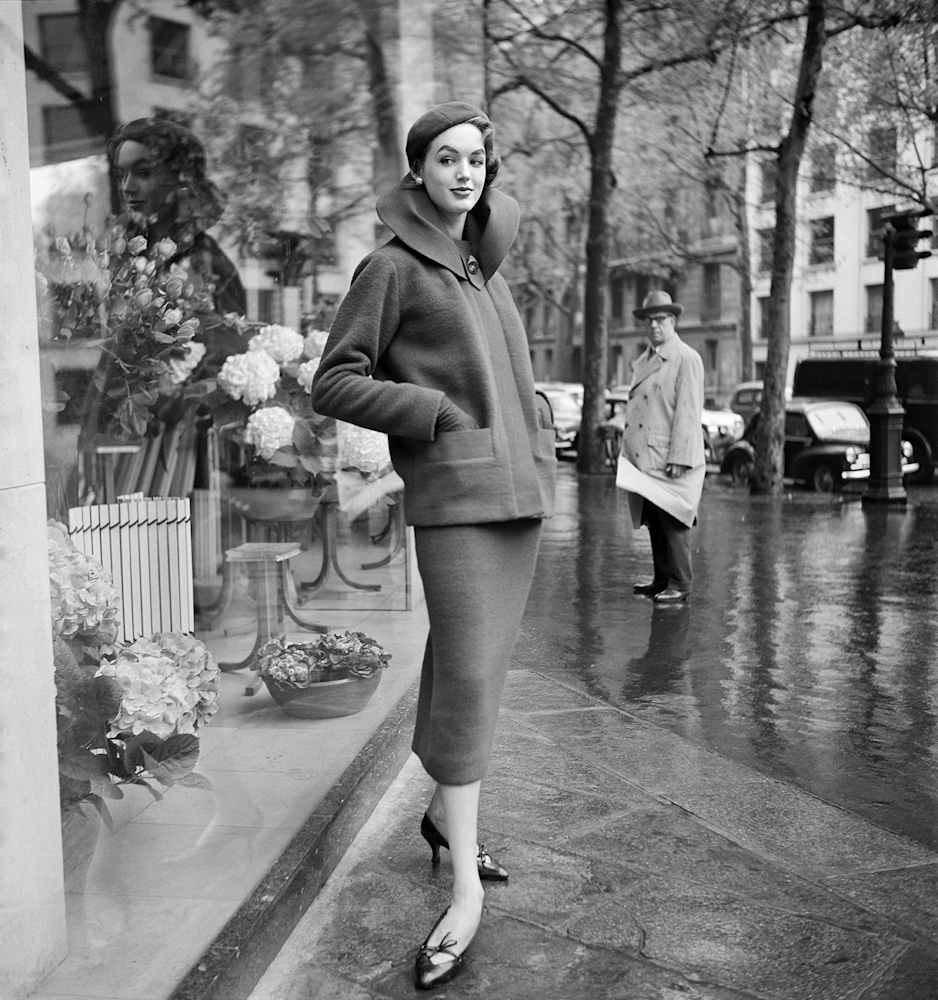 Black and white photo. A woman poses outside a shop in a fashionable 1950s outfit. A man in the background of the image looks towards her.