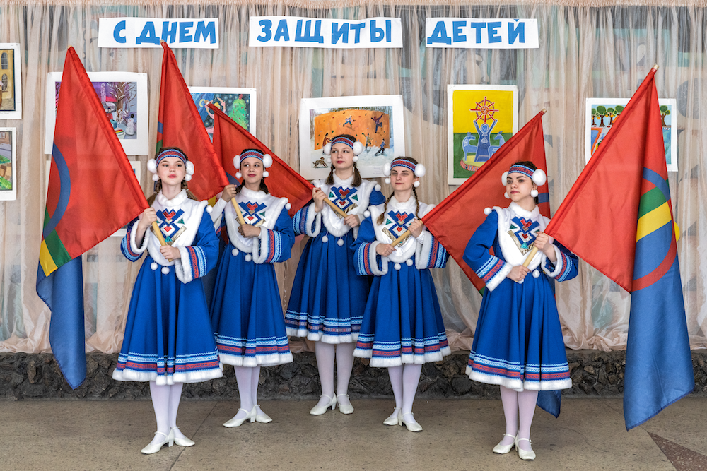 Women wearing traditional Saami clothing - blue dresses with white patterned shawls and headbands - while holding red flags.