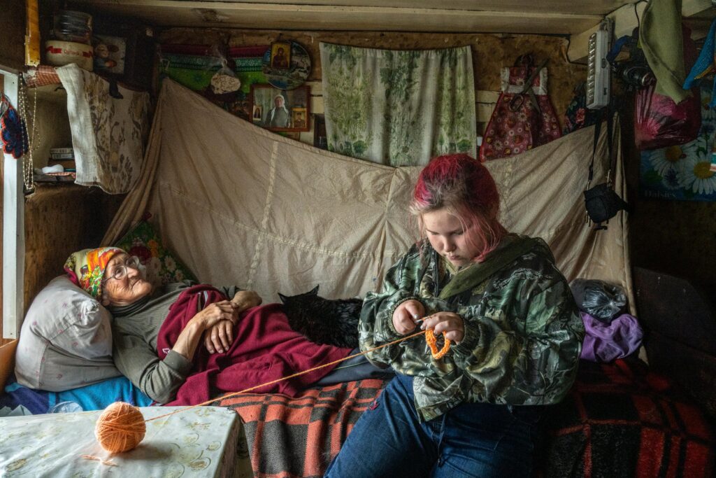 Uliana is knitting a decoration while an older woman lies on a bed behind her.