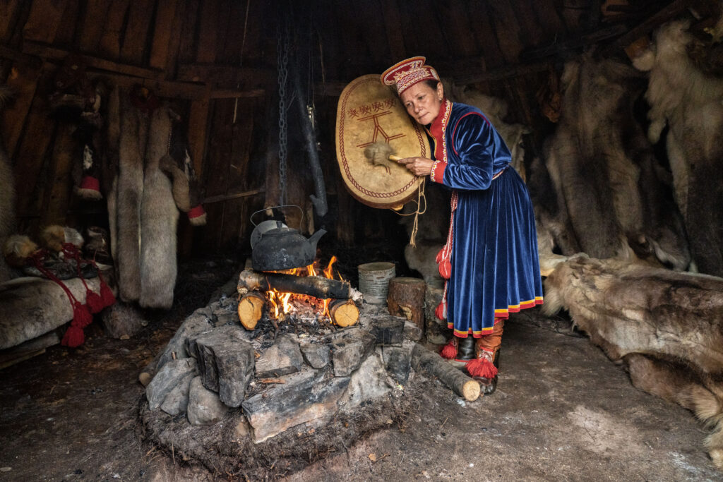 Elena Yakovleva, Uliana's aunt, prepares her drum to sound it and perform a ritual in front of a sacred stone in the Saami mini-village that she has reconstructed for tourists. She leans towards a small fire while holding the drum aloft.