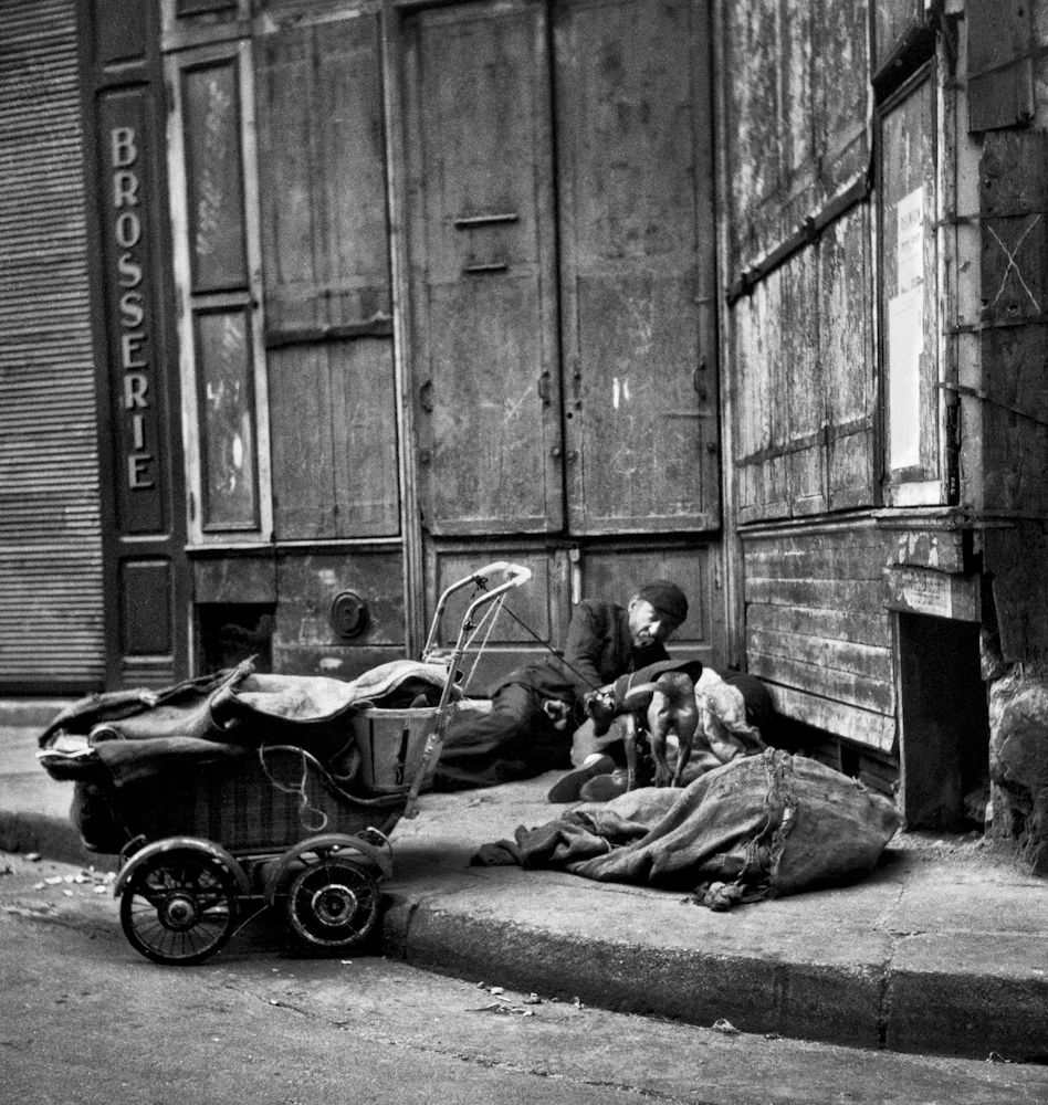 Black and white photo. A man, possible several people, are sleeping on a street. A dog stands over them.