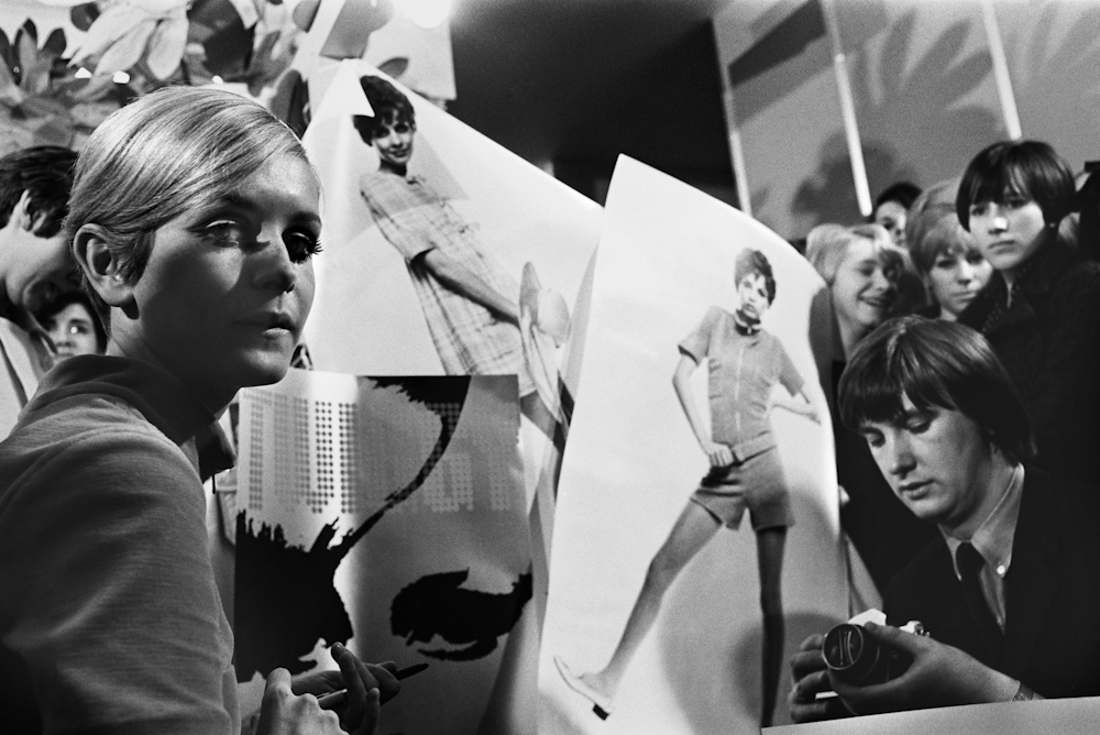 Black and white photo. A woman (Twiggy) looks towards the camera while a press photographer readies their camera.