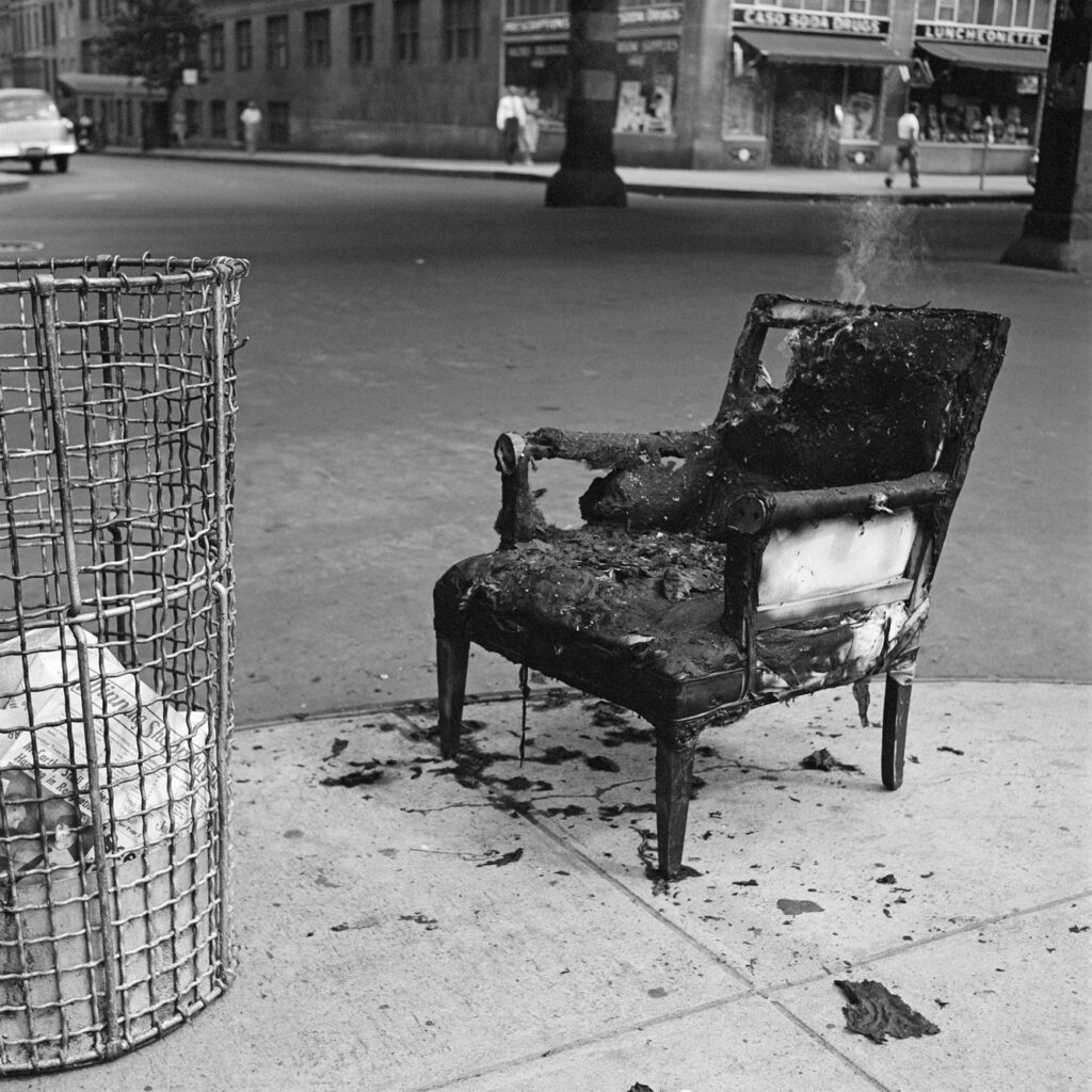 Black and white photo. A burned chair, still smoking, next to a bin.