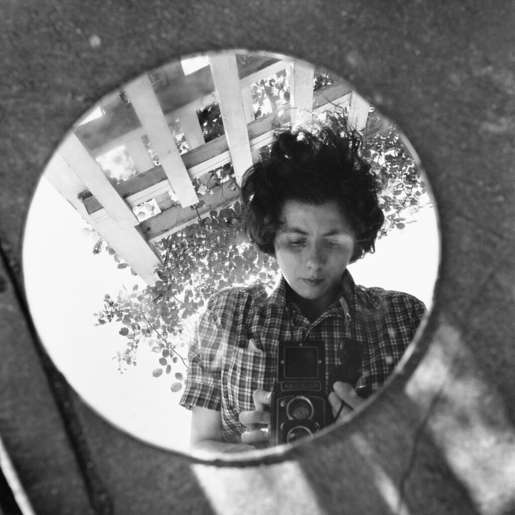 Black and white self portrait of Vivian Maier holding her camera, reflected in a circular mirror.