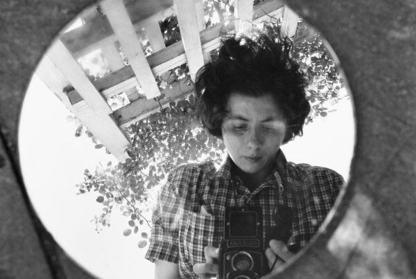 Black and white self portrait of Vivian Maier holding her camera, reflected in a circular mirror.