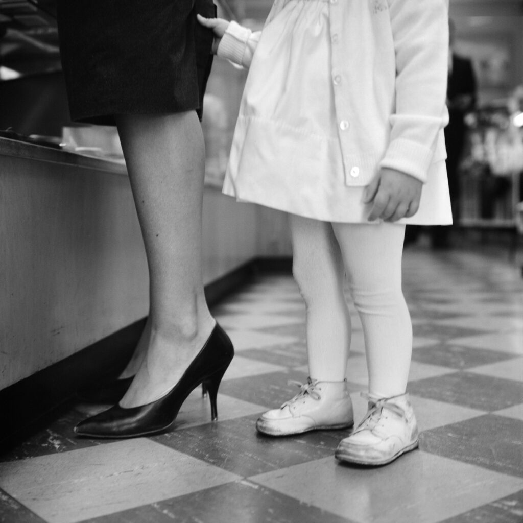 Black and white photo showing the legs and feet of a woman and a young girl standing at a shop counter.