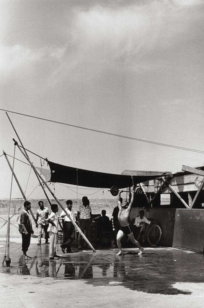 Black and white photo. A weight lifter performs a lift while a small crows looks on.