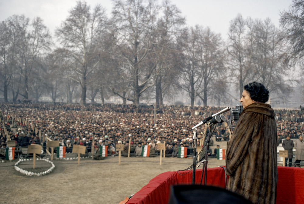 Colour photograph. Indira Gandhi stands at a microphone in front of a large crowd.