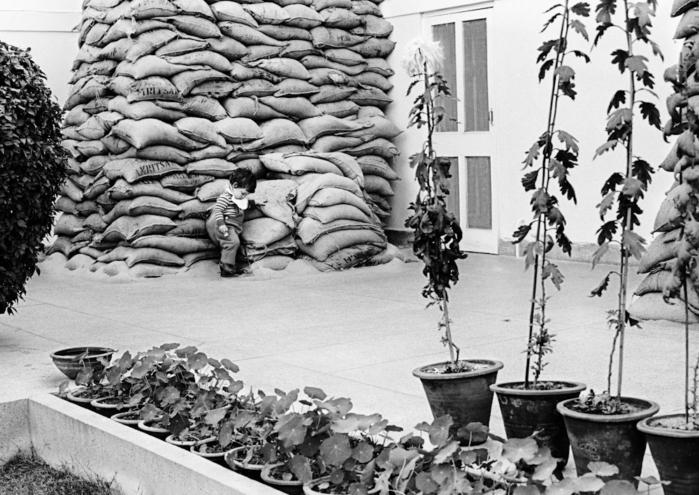 Black and white photo. A young boy plays with sandbags outside a house.