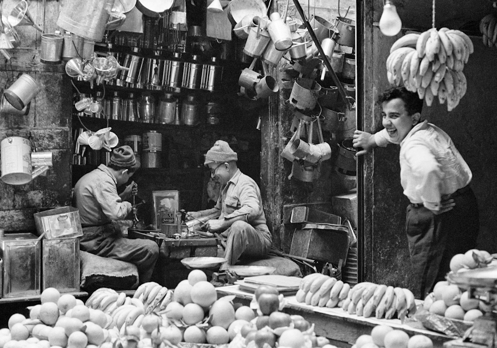 Black and white photograph of three men at a market stall in Lebanon, 1960. Two are seated, while a third stands and looks towards the camera. There are watering cans and fruit around them.