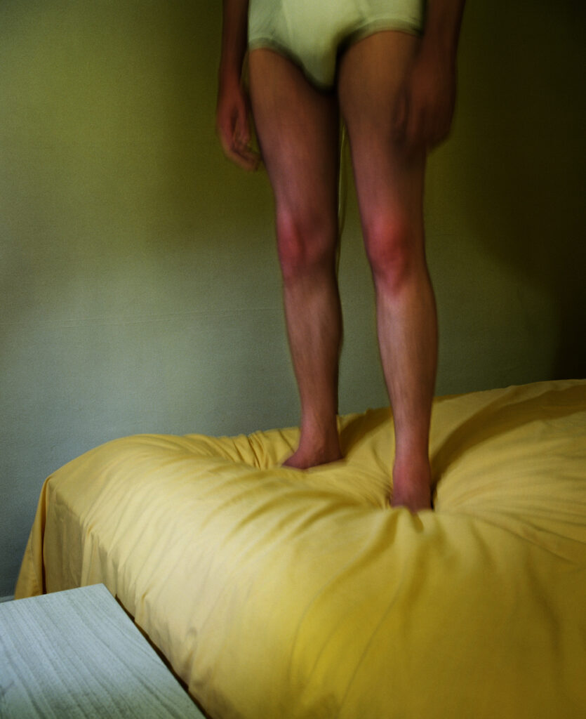 Two legs stand on a bed, covered with a yellow bed sheet.